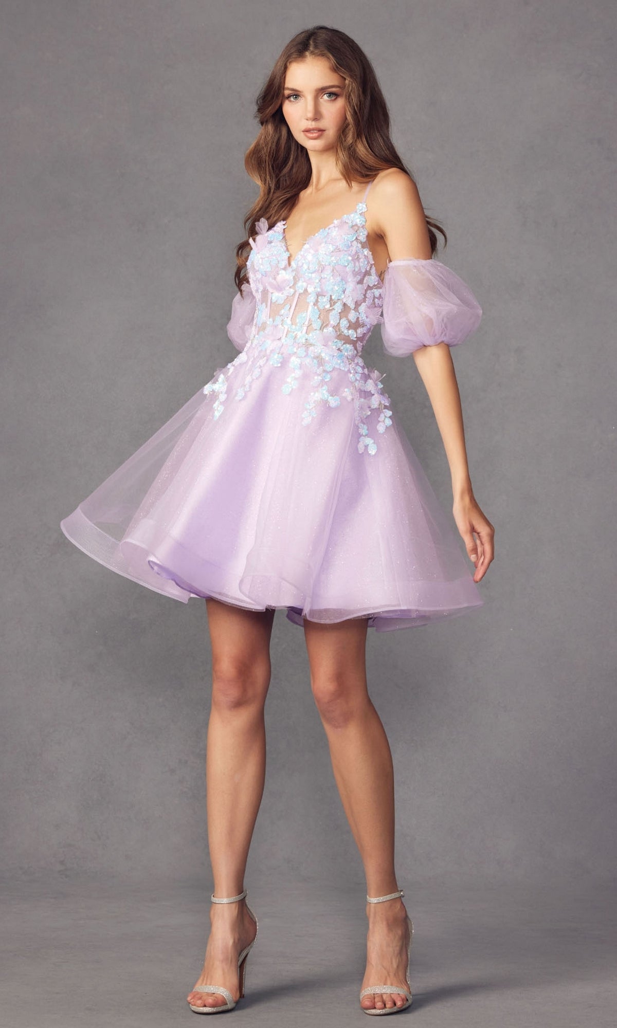 A woman poses against a gray background in the Puff-Sleeve Short Butterfly Homecoming Dress 901, a light purple babydoll style with sheer puff sleeves, floral appliqué on the bodice, and clear high-heeled sandals.